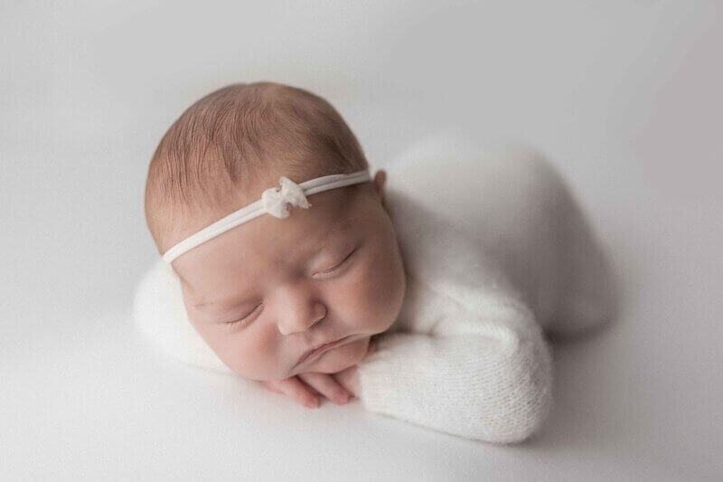 A newborn baby girl sleeps peacefully in a soft white outfit and matching headband, posed on a white backdrop during a studio photoshoot.