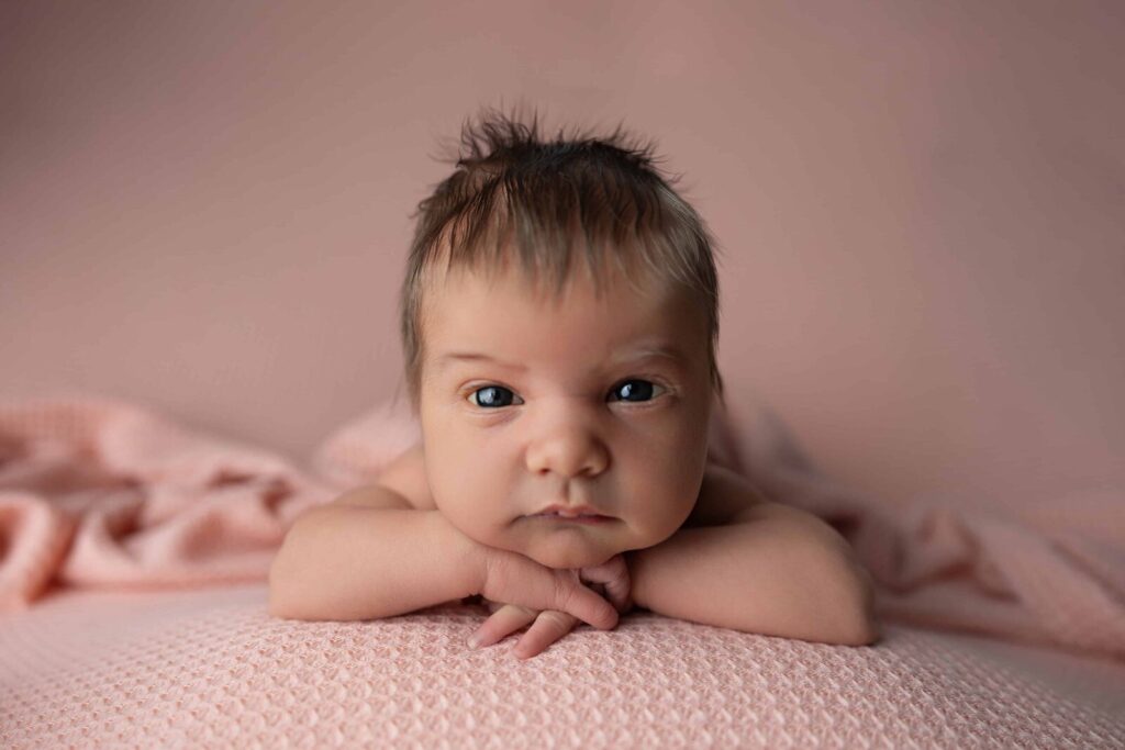 A newborn baby rests with their head on their hands, gazing toward the camera while lying on a soft pink textured blanket in a studio setting.