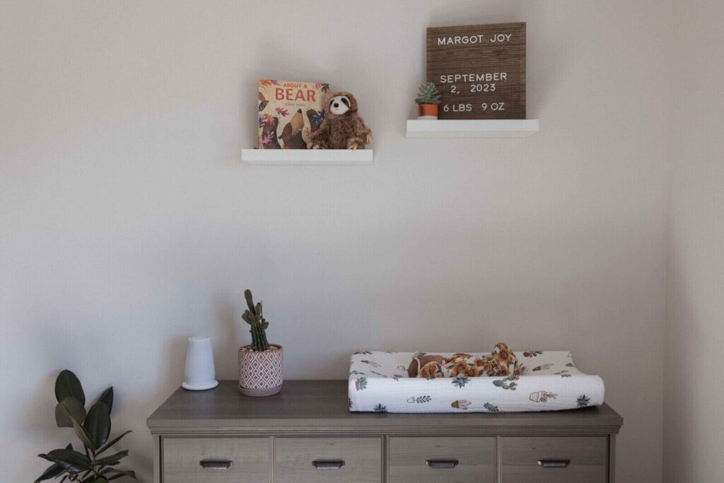 A newborn baby lies in a sunflower-patterned outfit on a changing pad in a nursery, with a name sign and baby book displayed on the wall above.