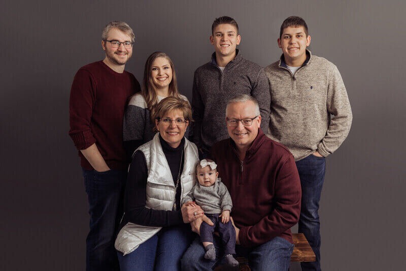 A family of seven, including a baby, poses together in a studio setting with coordinated casual outfits and neutral tones.