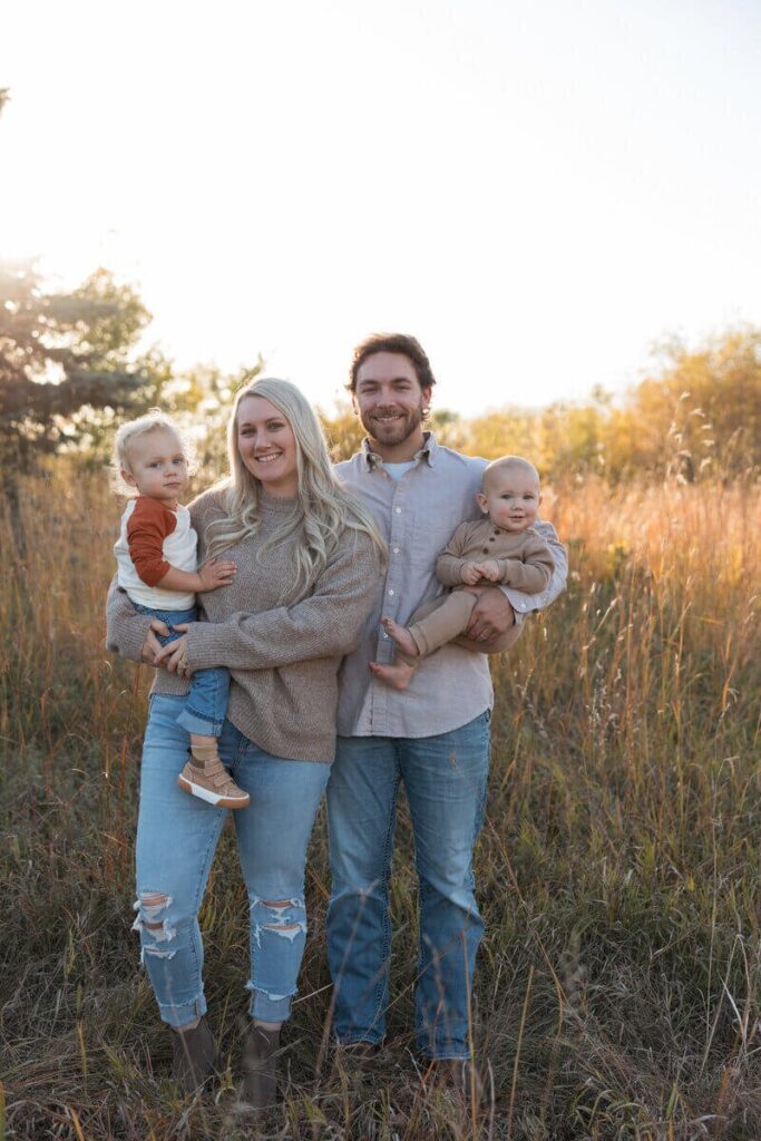A young family of four stands smiling in a sunlit field, each parent holding a child during a fall photoshoot.