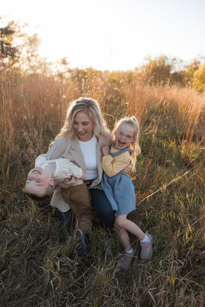 A mother laughs with her two children while playing together in tall golden grass during an outdoor session.