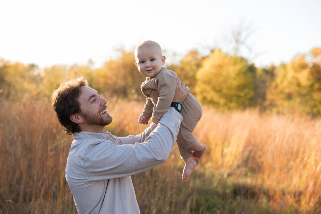 A father joyfully lifts his baby in the air during a golden hour photoshoot in a field of tall grass and autumn trees.