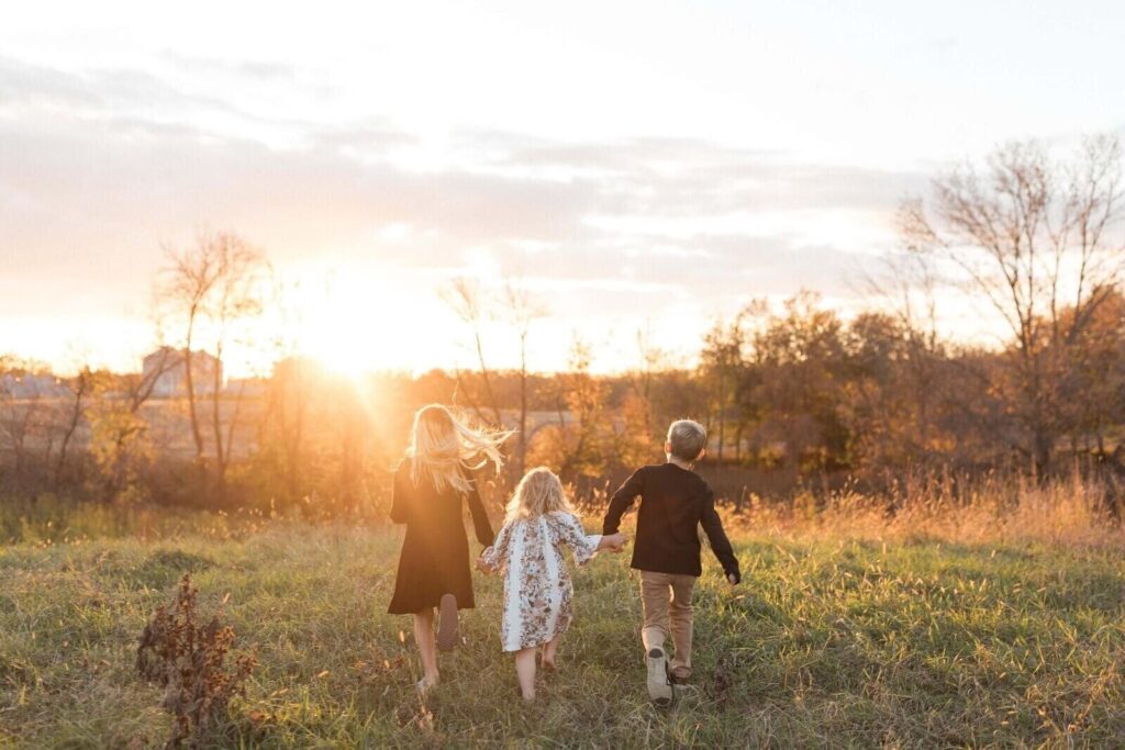 Three children holding hands run barefoot through a grassy field at sunset during a golden hour photoshoot.