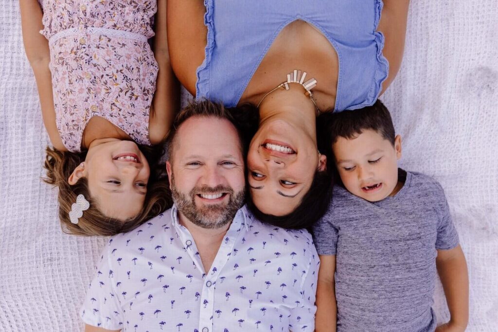 A family of four lies side by side on a white blanket, smiling and looking at each other during an outdoor photoshoot.
