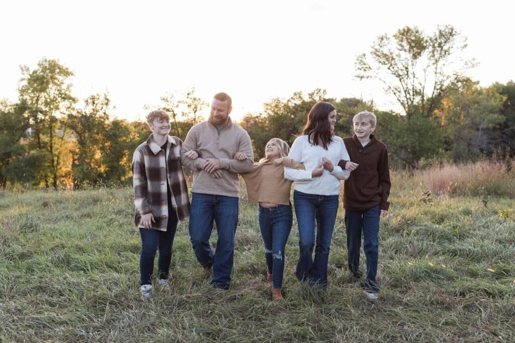 A family of five walks arm-in-arm through a grassy field at sunset, smiling and enjoying a candid moment together.