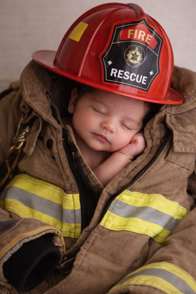 Newborn baby dressed in firefighter jacket and helmet sleeping.