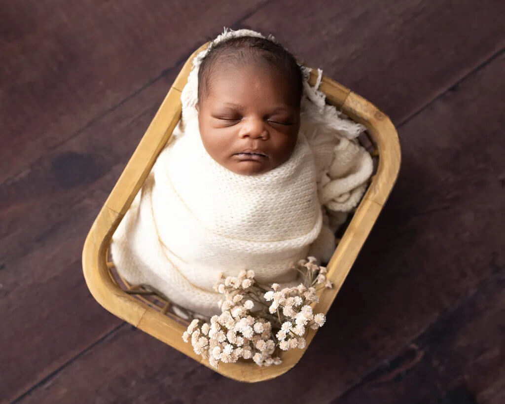 Newborn baby wrapped in a soft cream blanket sleeping in a wooden basket with delicate dried flowers