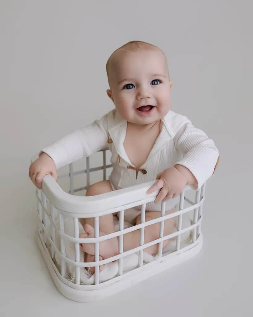 Smiling baby sitting in a white basket wearing a soft neutral outfit during a sitter session