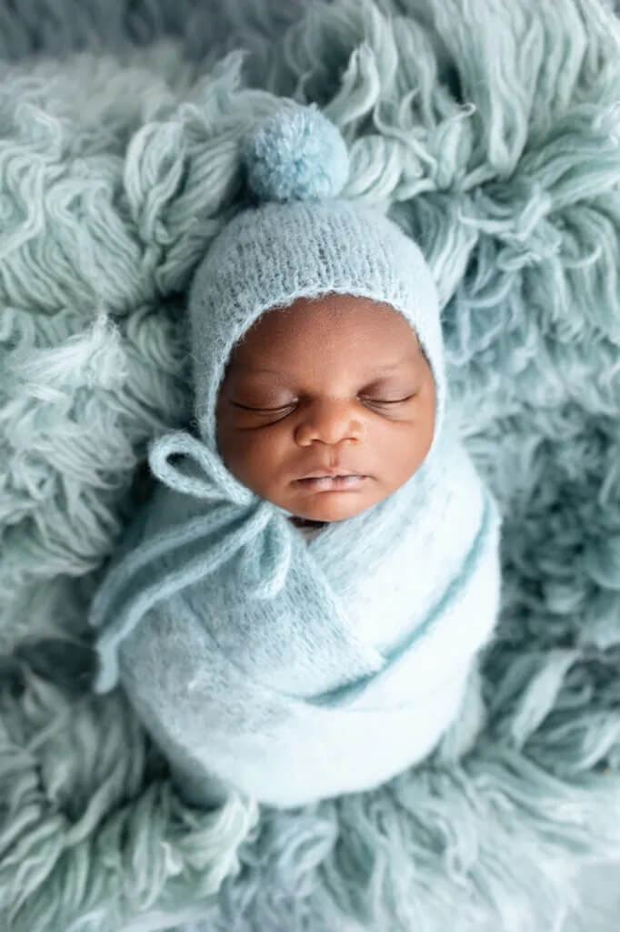 Newborn baby wrapped in a soft blue swaddle lying on a textured fluffy rug wearing a matching bonnet