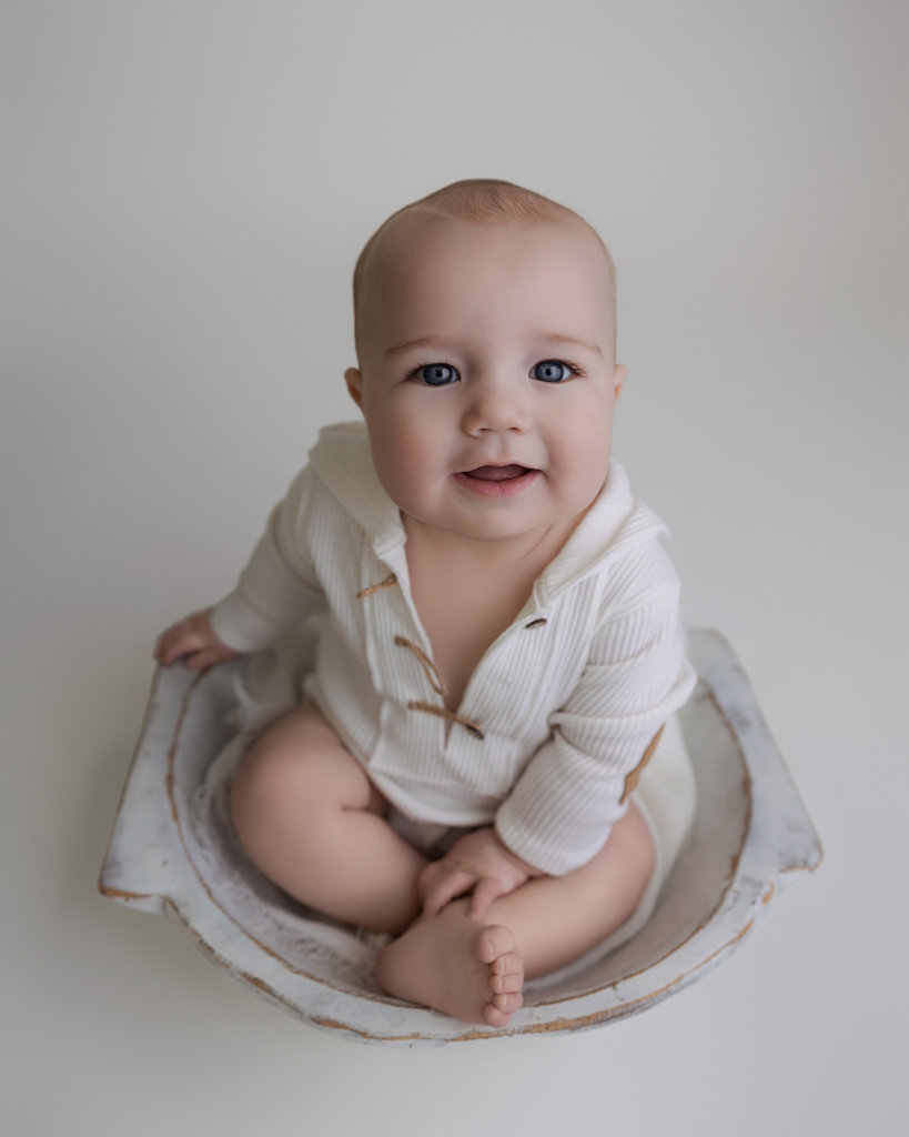 Happy baby sitting in a white bowl prop wearing a neutral outfit and smiling at the camera
