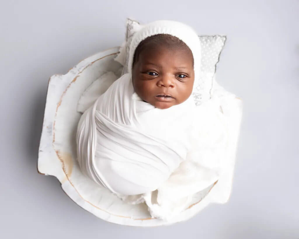 Newborn baby wrapped in a white swaddle posed in a white bowl prop looking awake