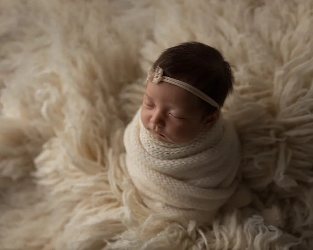 Newborn baby girl wrapped in a cream knit blanket lying on a soft fluffy rug wearing a small headband