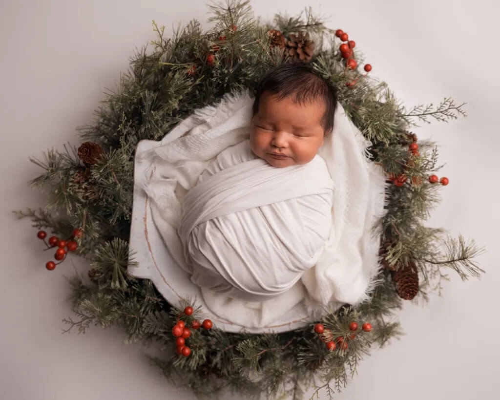 Newborn baby wrapped in a white blanket posed inside a Christmas wreath with pinecones and red berries