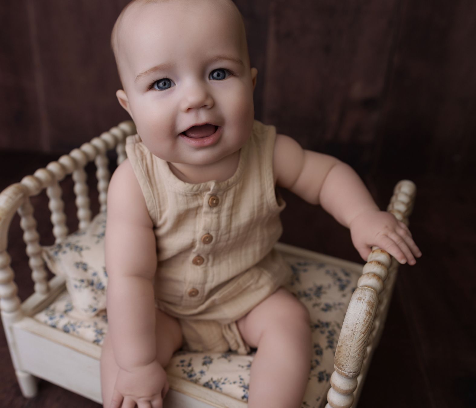 Smiling baby sits on dolls bed in brown baby romper