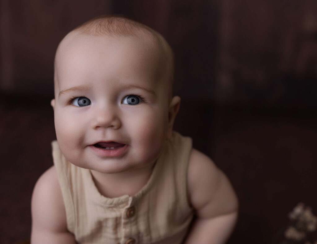 Baby smiling during a sitter session with a dark wooden backdrop and neutral outfit.