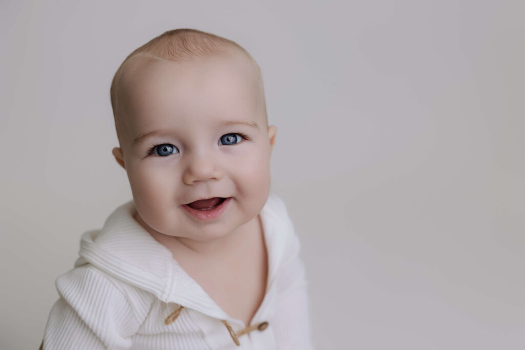 Close-up portrait of a smiling baby boy in a white outfit against a light studio background.