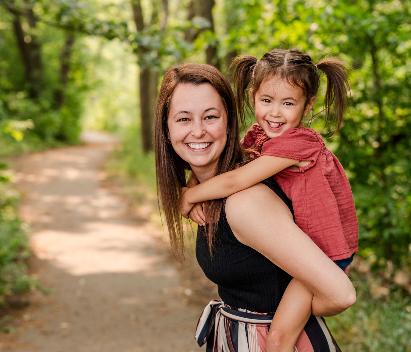 Melissa Sampson holding her young daughter on her back in the sunny outdoor park.