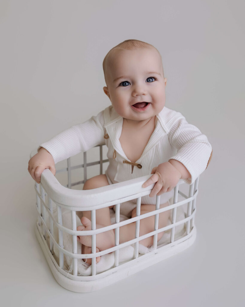 Baby sitting in a white basket wearing a white outfit in a bright studio portrait.