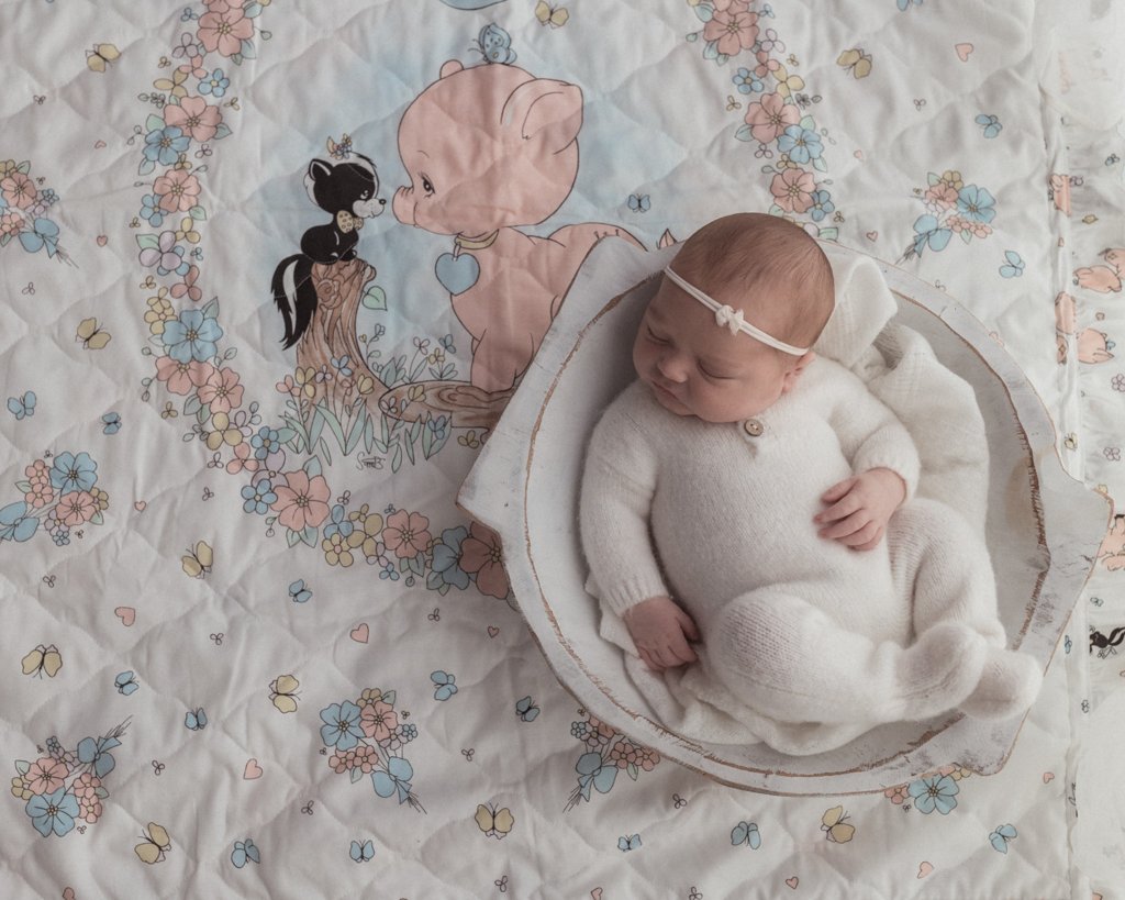 A sleeping baby laying in a wooden bowl on top of an old blanket featuring a pig and a skunk. 