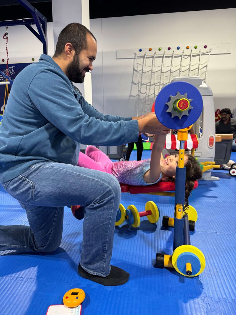 Man helping a child play on toy fitness equipment inside We Rock the Spectrum Kid's Gym sensory gym and indoor play center.