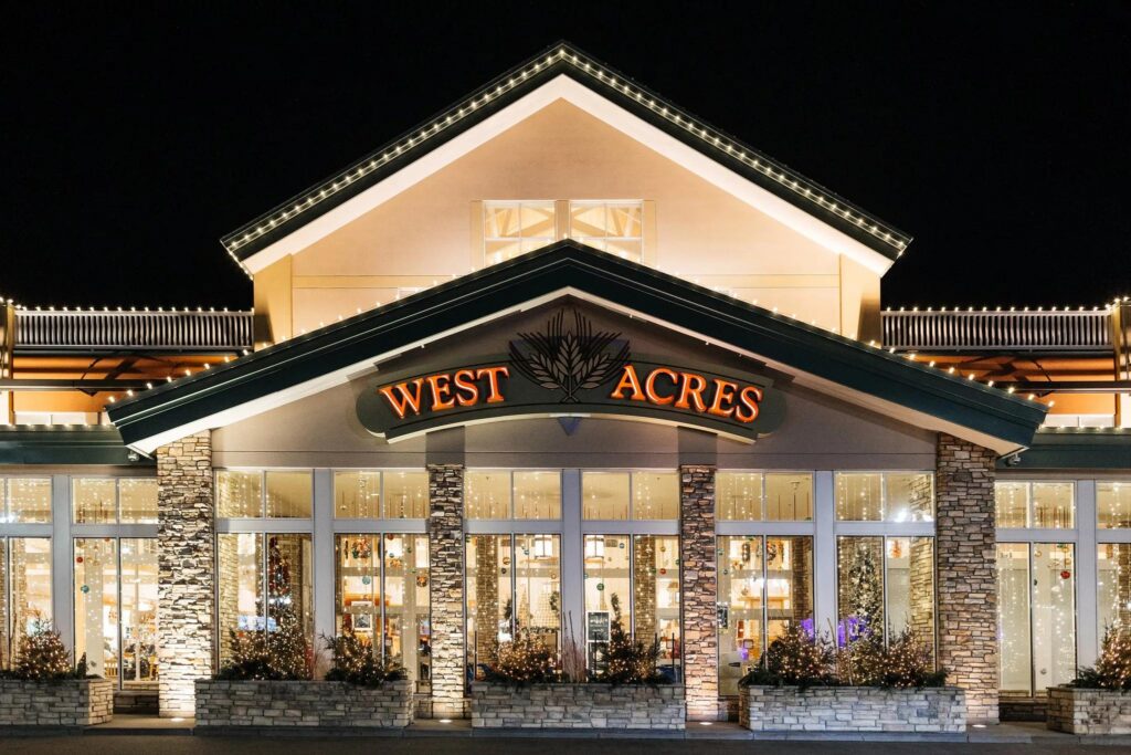 Exterior entrance of West Acres Mall lit up at night with holiday lights and storefront displays.